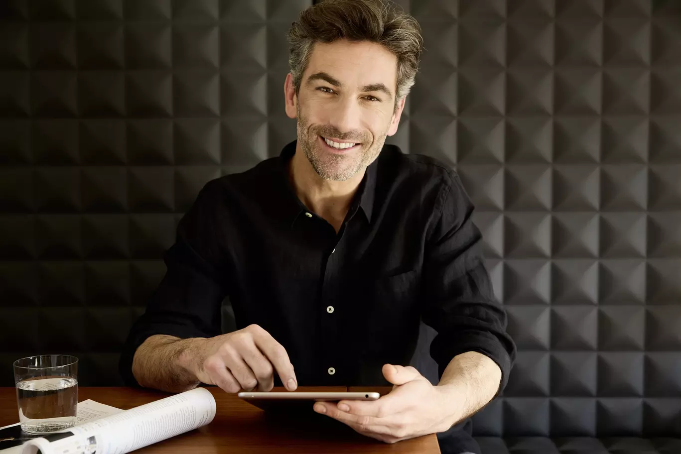 Mann mittleren Alter, der in entspannter Atmosphäre mit einem Tablet in der Hand vor einem dunklen Hintergrund sitzt, symbolisiert den Kundenservice von Alpecin.