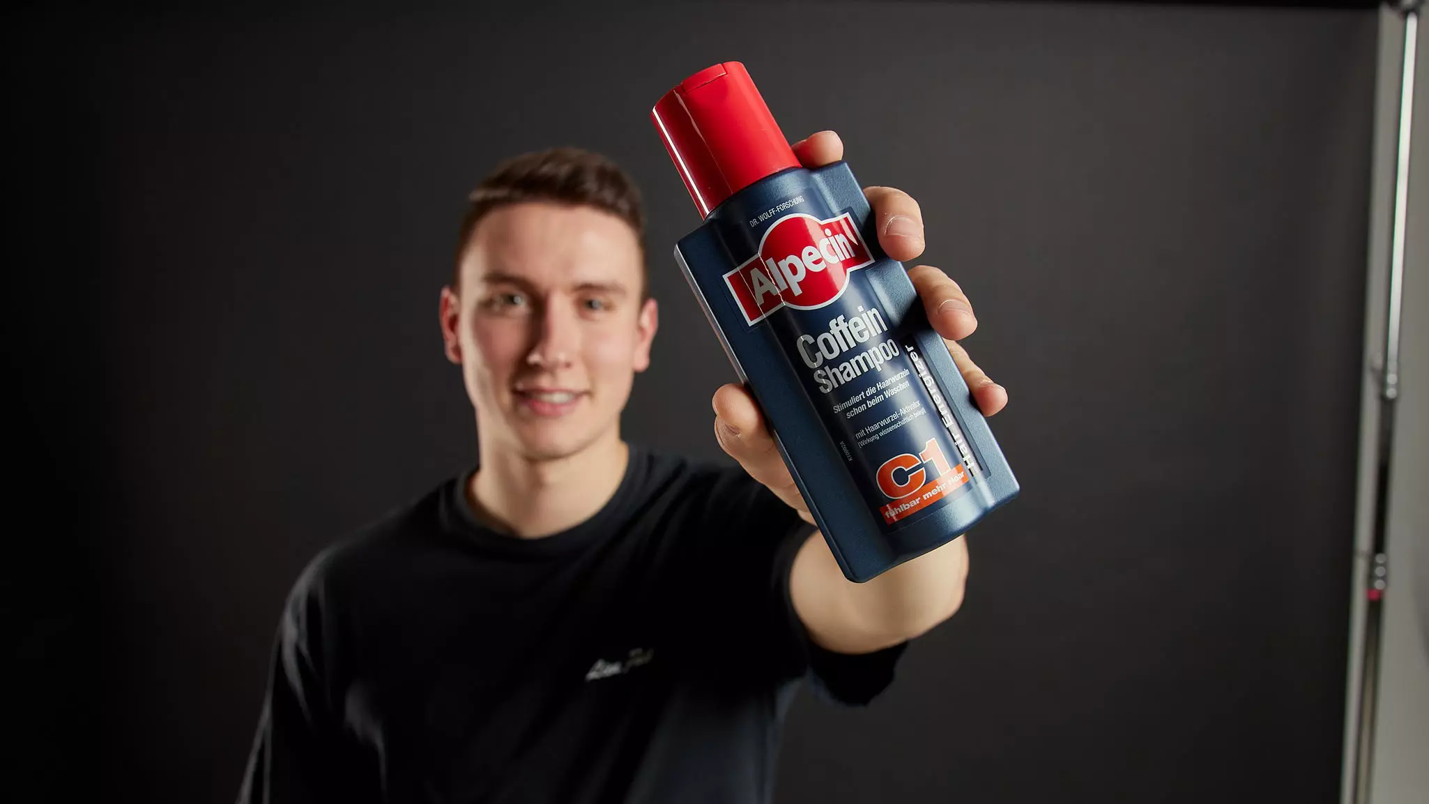 Eine junger Mann hält eine blaue Flasche mit rotem Deckel, beschriftet mit "Alpecin Caffeine Shampoo C1." Er trägt ein schwarzes Hemd vor einem dunklen Hintergrund und präsentiert das Produkt.