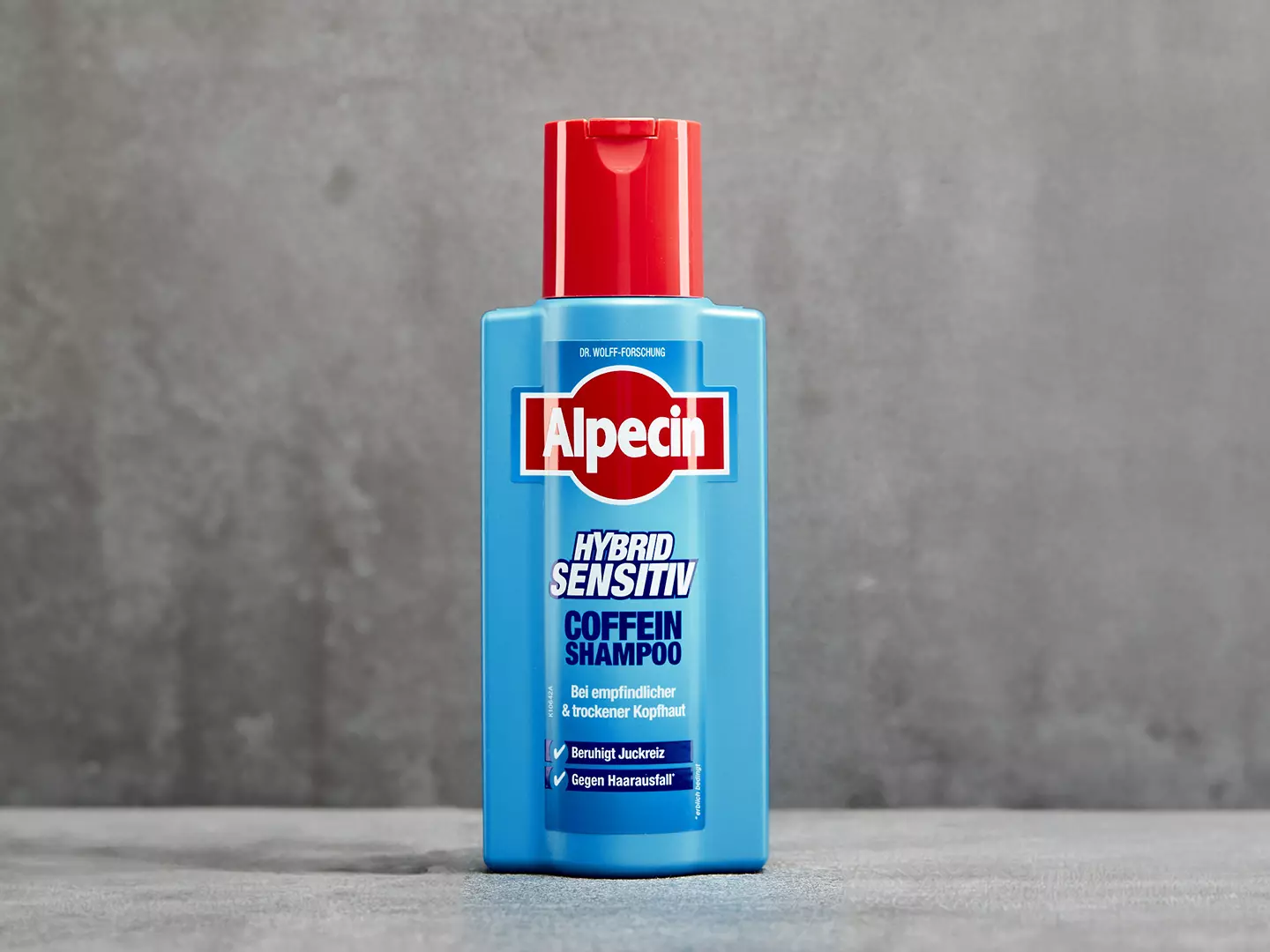 Eine hellblaue Flasche Alpecin Hybrid Sensitiv Shampoo steht auf einer grauen Fläche, um die Aussage zu unterstützen, dass ein mildes, feuchtigkeitsspendendes Shampoo bei Juckreiz aufgrund einer trockenen Kopfhaut helfen kann