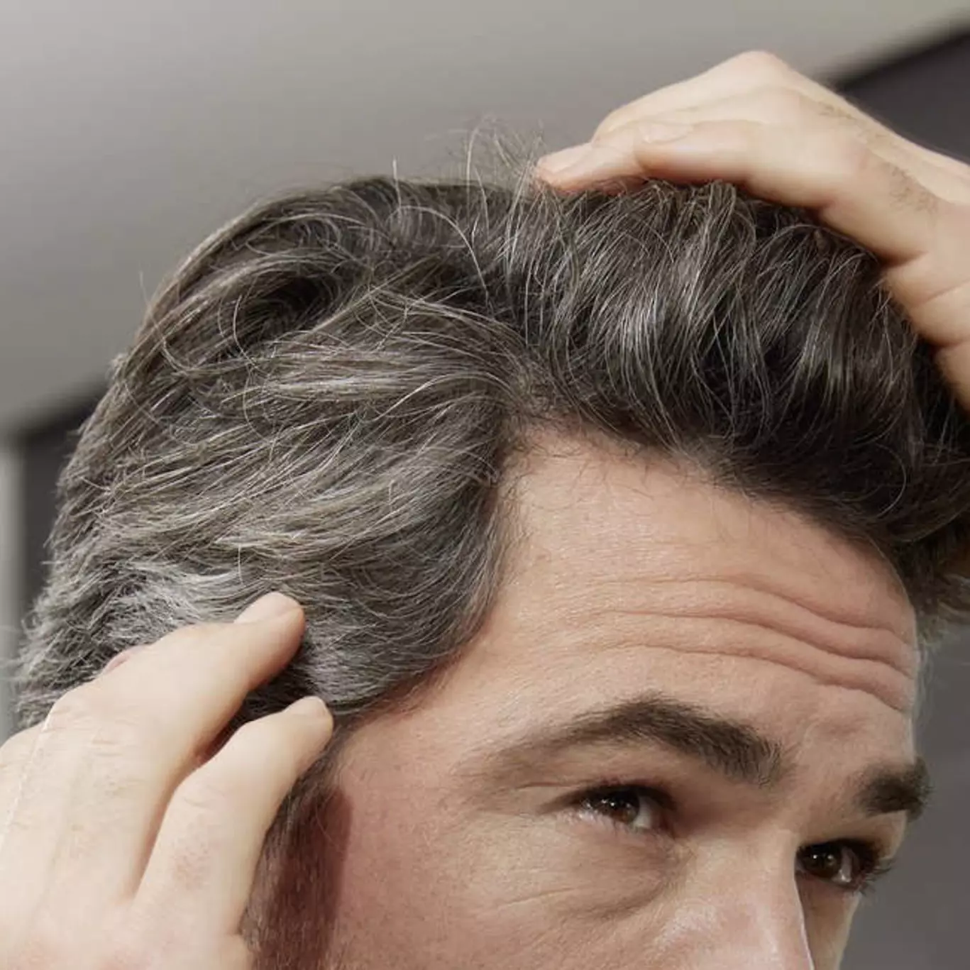 Ein Mann berührt mit beiden Händen sein ergrauendes Haar, fokussiert auf die Haarstruktur und Farbe, in einer modernen Innenumgebung.
