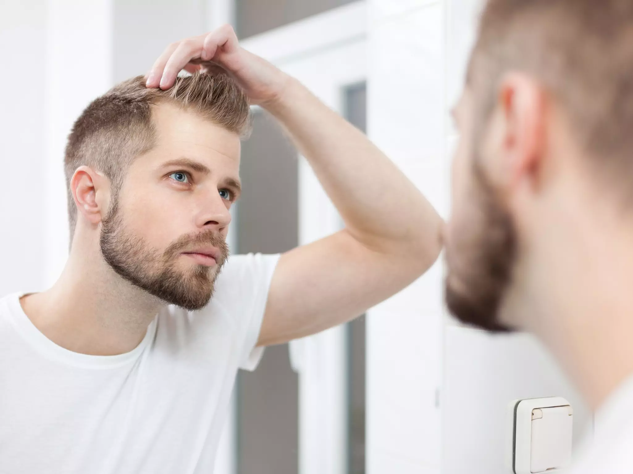 Ein junger Mann steht vor einem Spiegel und betrachtet seine Geheimratsecken als Sinnbild für die Frage "Wie viel Haarausfall ist normal?"