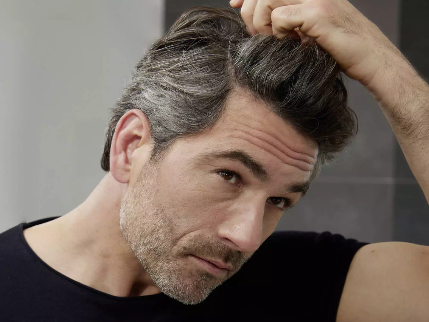 Mann mittleren Alters mit grauen Haaren an den Schläfen aber noch dunklem Haupthaar greift sich ins Haar und begutachtet kritisch seine grauen Haare